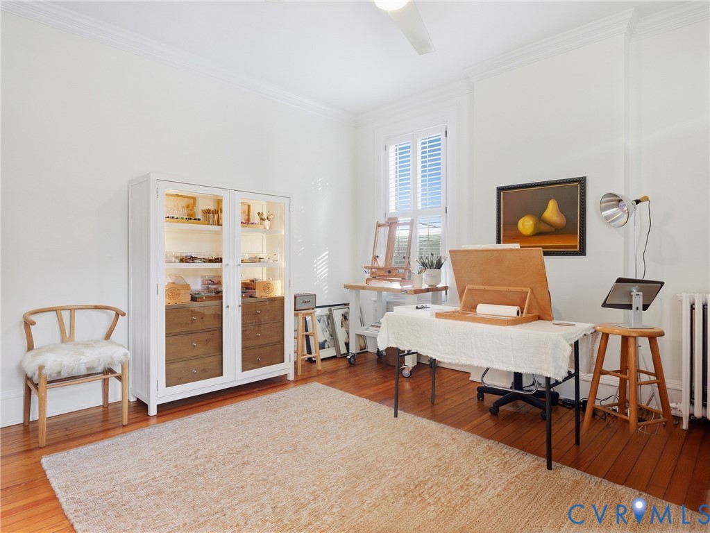 Hardwood flooring, white painted walls, crown molding, a window with louvered shutters, and a ceiling fan