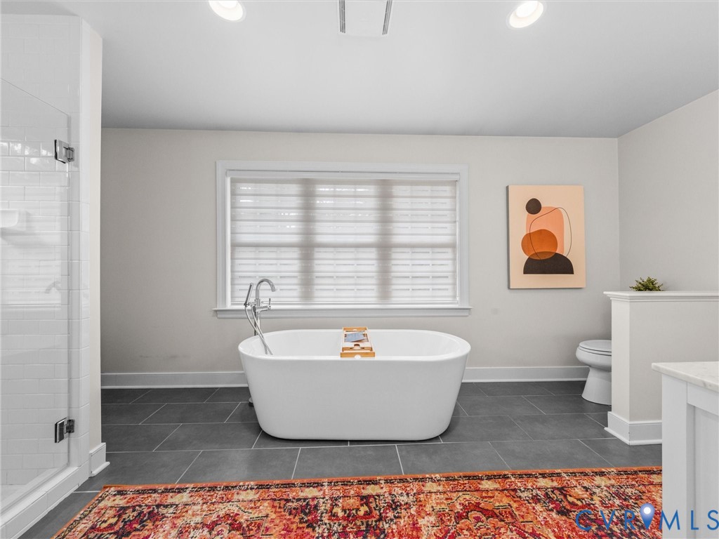 Freestanding soaking tub with floor-mounted faucet, walk-in shower with glass enclosure and subway tile, dark gray floor tiling, and recessed ceiling lighting