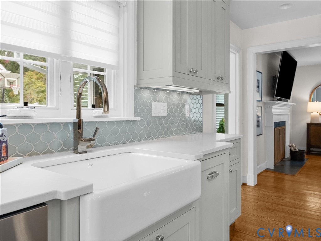 White farmhouse sink with a gooseneck faucet, light blue arabesque tile backsplash, light-toned cabinetry, white countertops, and hardwood flooring