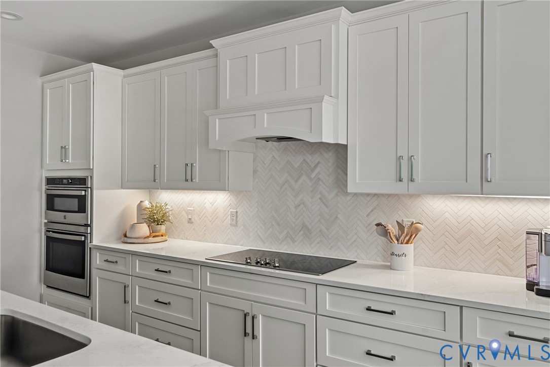 White shaker cabinetry with crown molding, integrated stainless steel double oven, herringbone tile backsplash, integrated cooktop, and undermount lighting