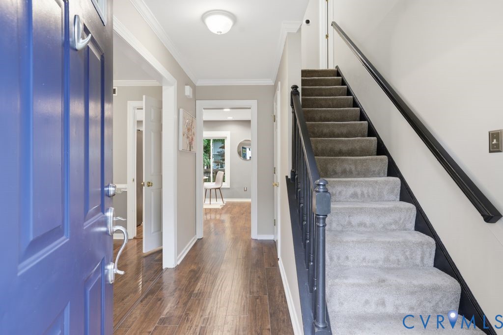Welcoming foyer featuring a striking cobalt blue entry door, wide-plank wood-finish flooring, and a graceful staircase with gray carpeted treads and black metal handrailing