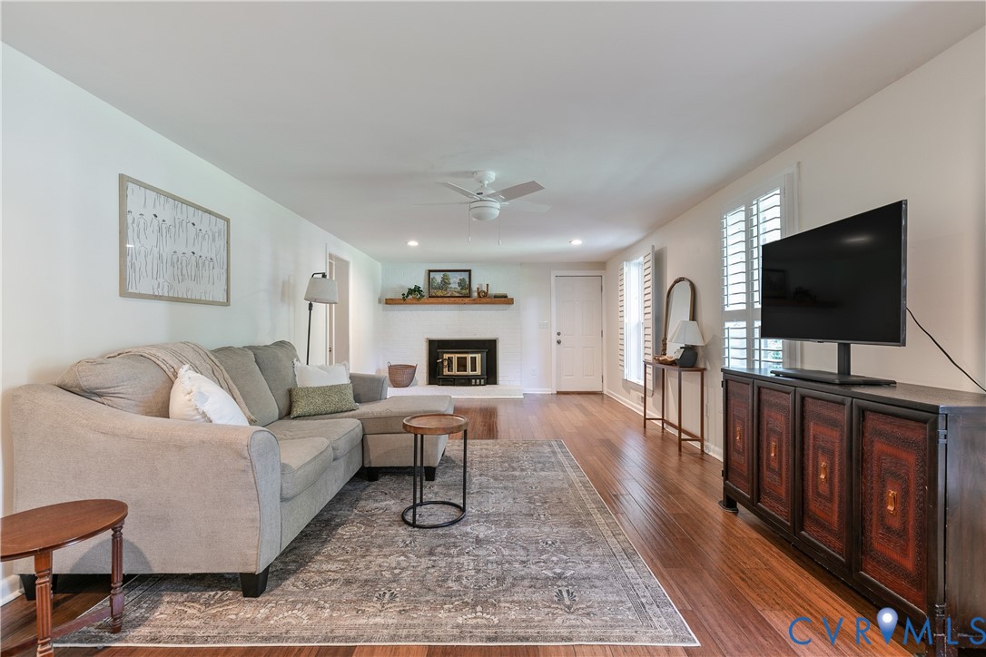 Spacious living area featuring wood-finish flooring, a white brick fireplace with a wood mantle, and white plantation shutters on the windows
