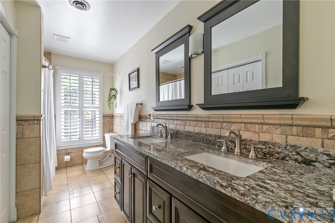 Dual sink vanity with stone countertop, tile flooring, and a window with plantation shutters