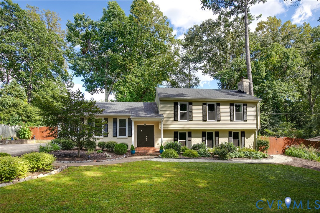 Split-level residence featuring a front entry, multiple windows with shutters, a brick chimney, and mature landscaping