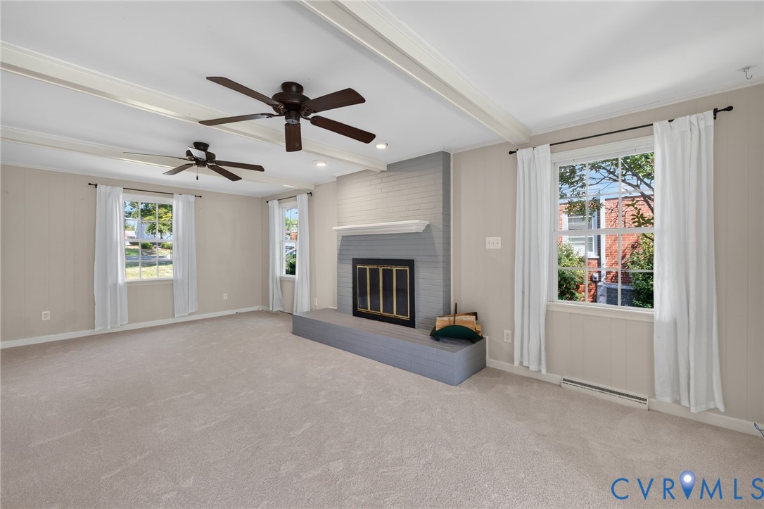 Spacious living area featuring exposed beam ceilings, two ceiling fans, a brick-surround fireplace with a mantel, and two windows
