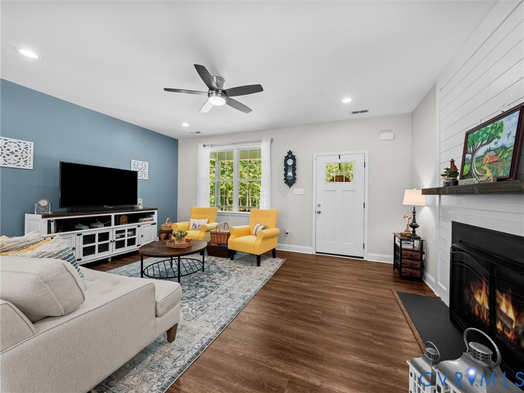 Expansive living area featuring wood-finish flooring, a ceiling fan with integrated lighting, a fireplace with a mantel, and a multi-pane window