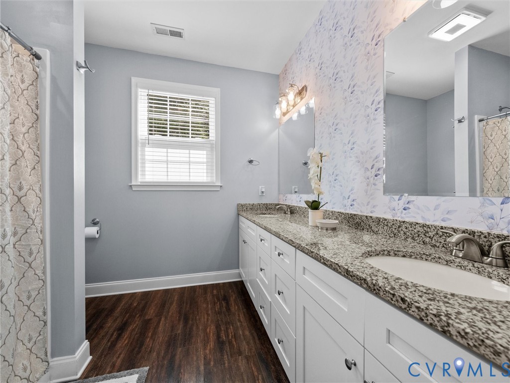 Double vanity with granite countertops, wood-finish flooring, two large mirrors, and a window with blinds