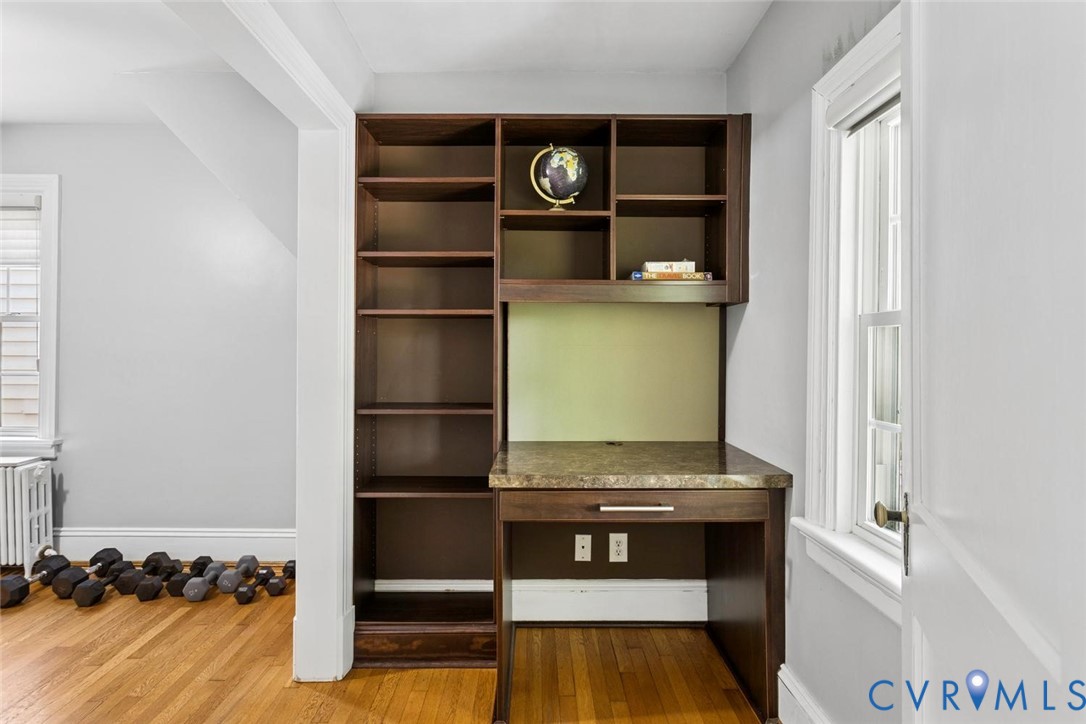 Built-in desk with a stone-finish countertop and integrated shelving, hardwood flooring, and a window with white trim