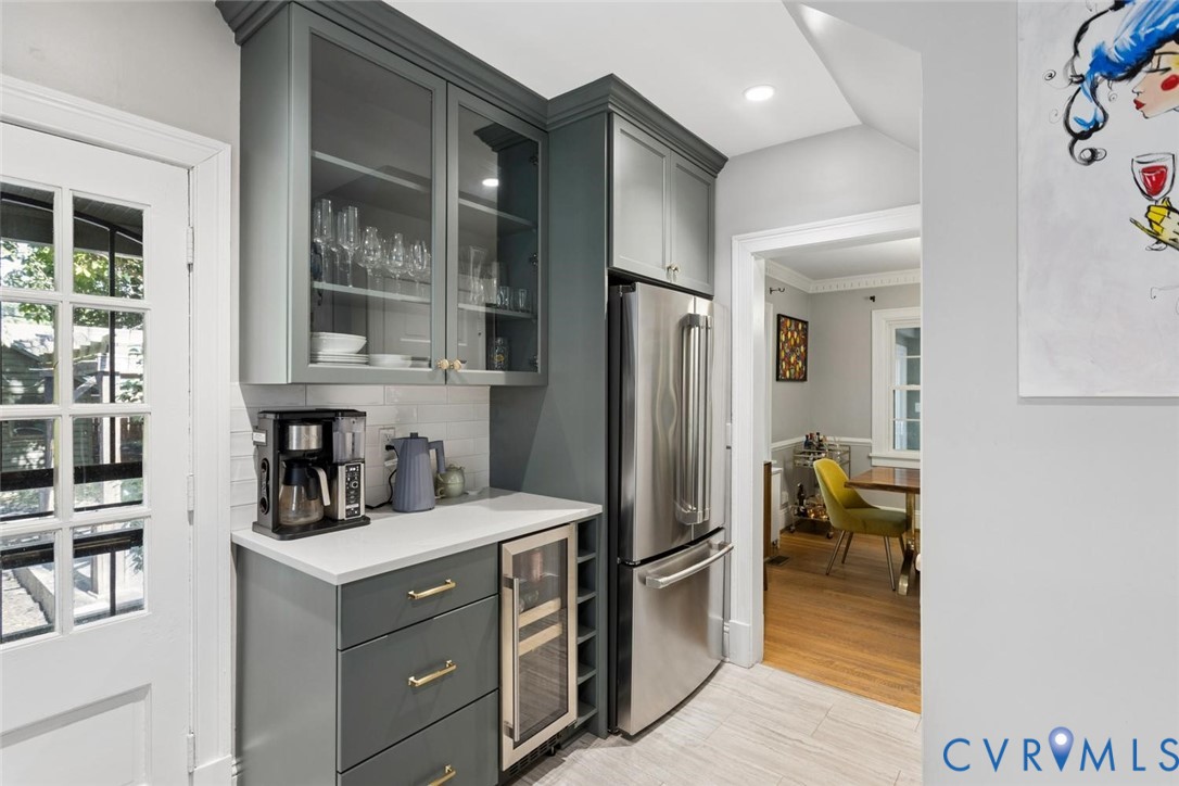 Integrated stainless steel refrigerator, built-in beverage cooler, white subway tile backsplash, light-toned countertops, and grey cabinetry with gold-toned hardware