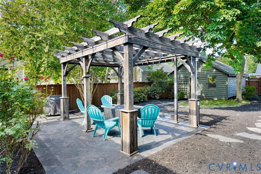 Wood pergola on a dark paver patio with a built-in outdoor speaker system, surrounded by mature trees and shrubs