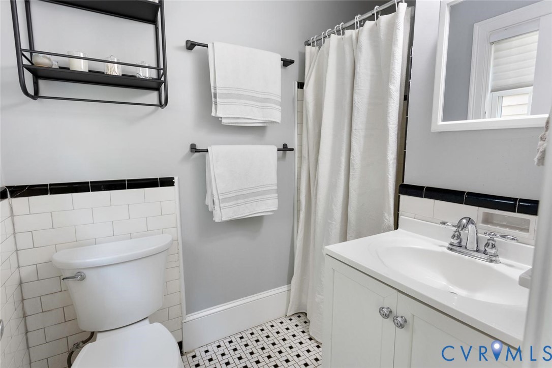 White subway tile wainscoting with black trim, black and white geometric tile flooring, white vanity with integrated sink, chrome fixtures, and a wall-mounted shelving unit