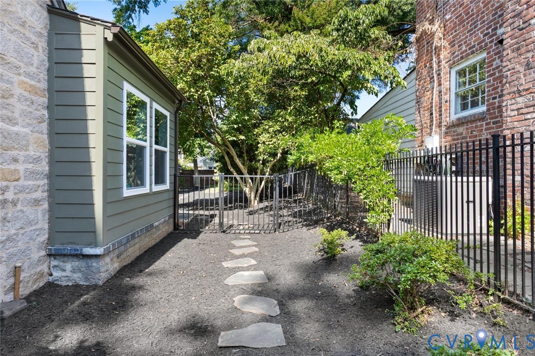 Stone and siding exterior with multiple windows, leading to an enclosed outdoor space featuring stepping stones, mature trees, and a black wrought iron fence