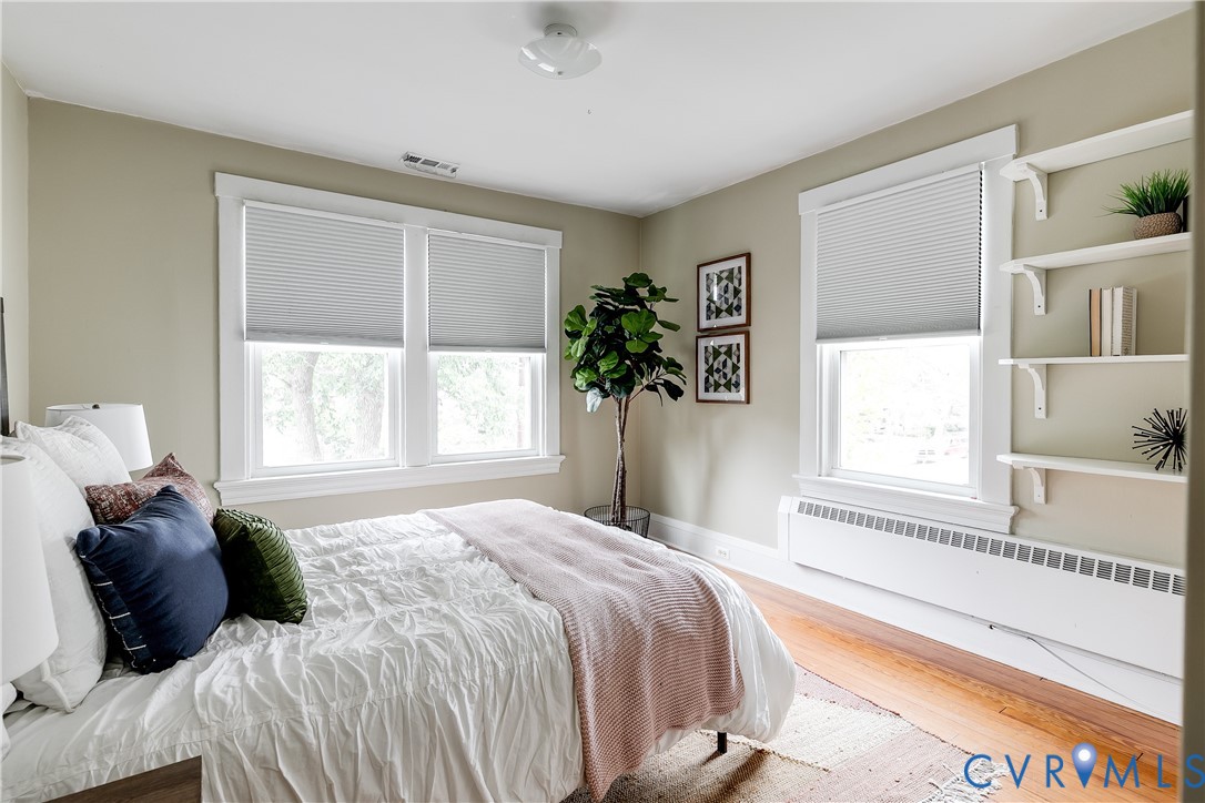 Bright, sun-filled bedroom with dual windows featuring cellular shades, hardwood flooring, and built-in shelving with display space
