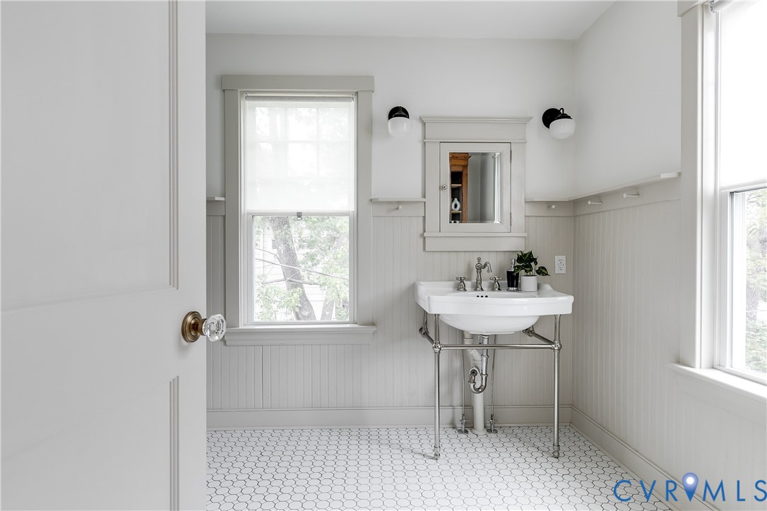 Pedestal sink with chrome-finish stand, integrated medicine cabinet with mirror, wall-mounted sconces, hexagonal floor tiling, and beadboard wainscoting