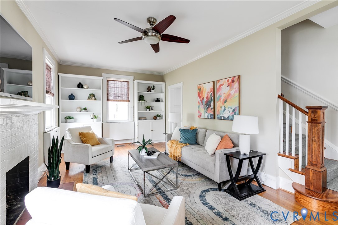 Hardwood flooring, white built-in shelving, white brick fireplace, wood staircase with white balusters, and a ceiling fan with wood-finish blades