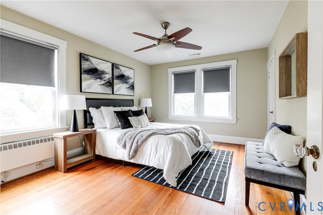 Hardwood flooring, painted walls, ceiling fan with light fixture, multiple windows with roller shades, and a built-in radiator