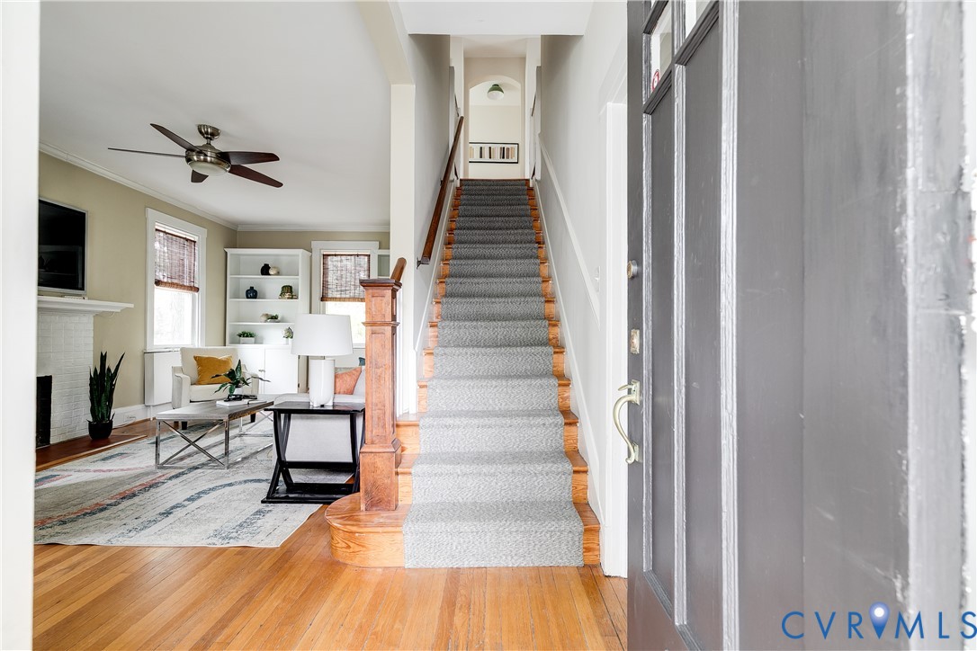 Warm wood flooring, a carpeted staircase with wood banister, built-in shelving, and a ceiling fan with integrated lighting