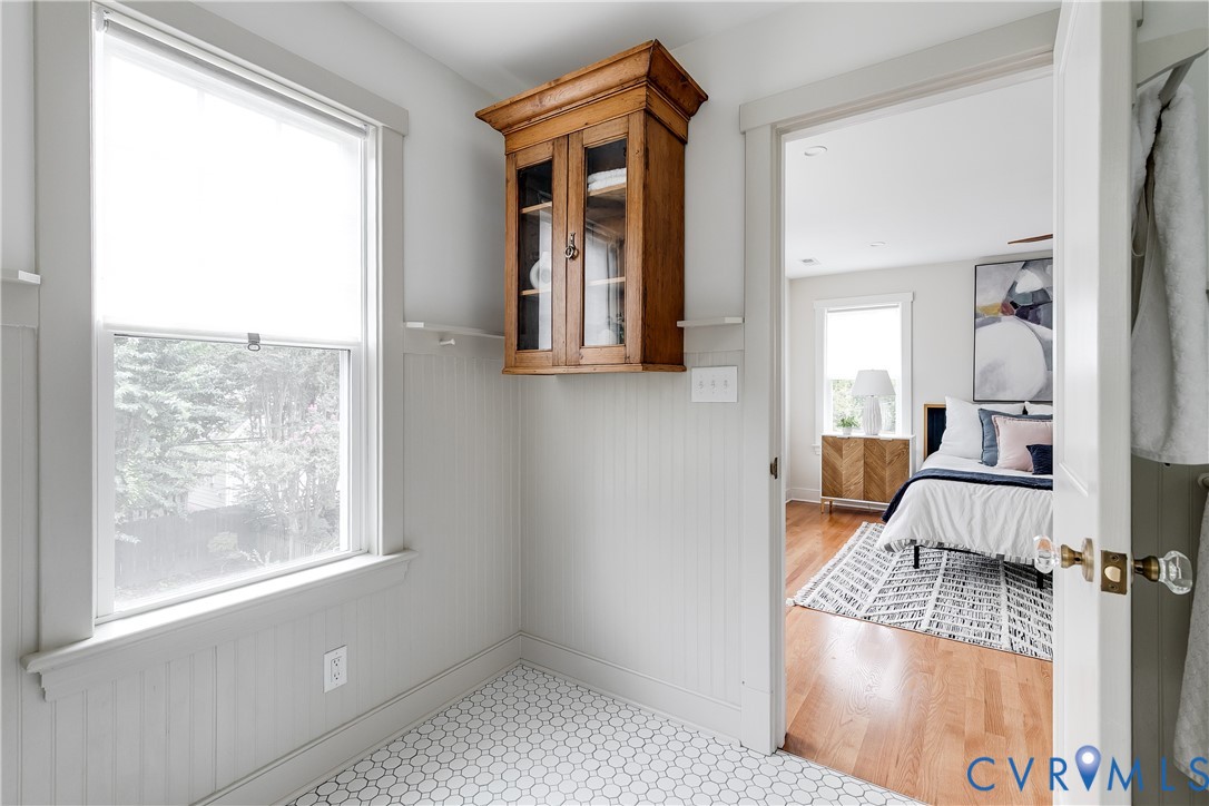 Hexagonal tile flooring, beadboard wall paneling, built-in wooden cabinetry, window with roller shade, and a door with clear glass knob