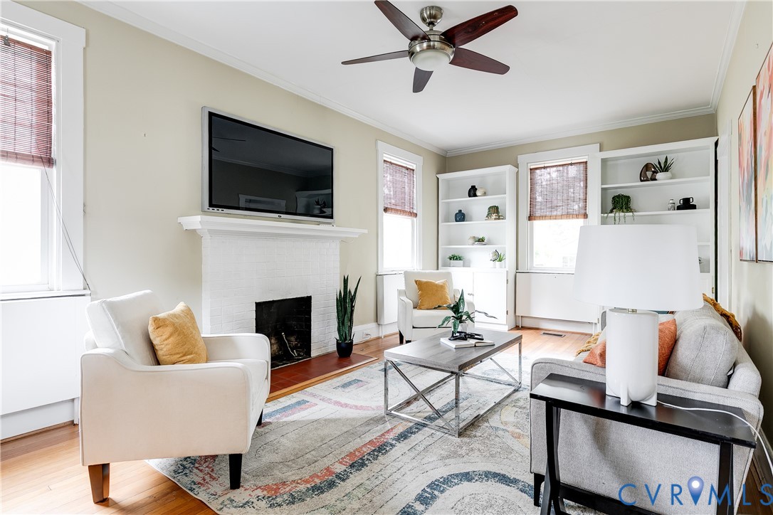 Hardwood flooring throughout, white painted brick fireplace with mantel, built-in shelving, ceiling fan with light fixture, and window treatments