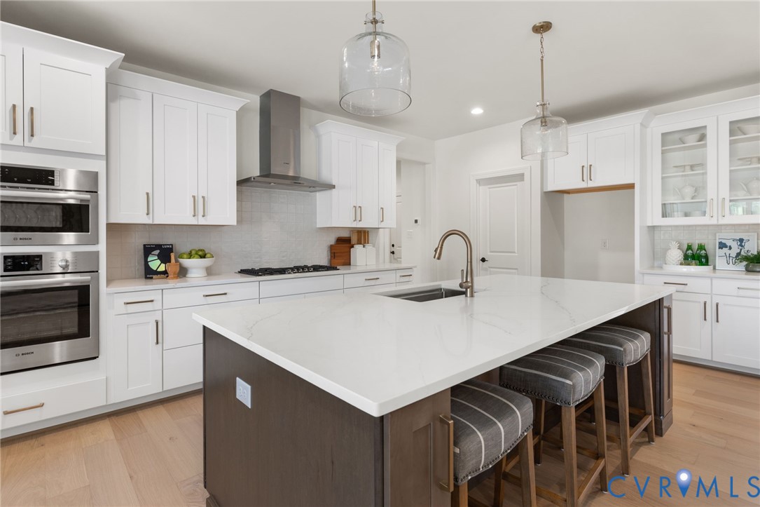 White cabinetry with brushed brass hardware, stainless steel built-in appliances, a central island with an integrated sink, pendant lighting, and wood-finish flooring