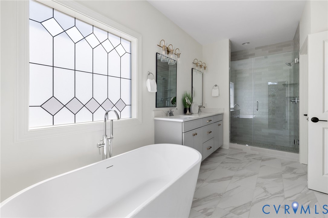Freestanding soaking tub with a floor-mounted faucet, dual vanity with undermount sinks, frameless glass-enclosed shower, geometric patterned privacy window, and large format tile flooring