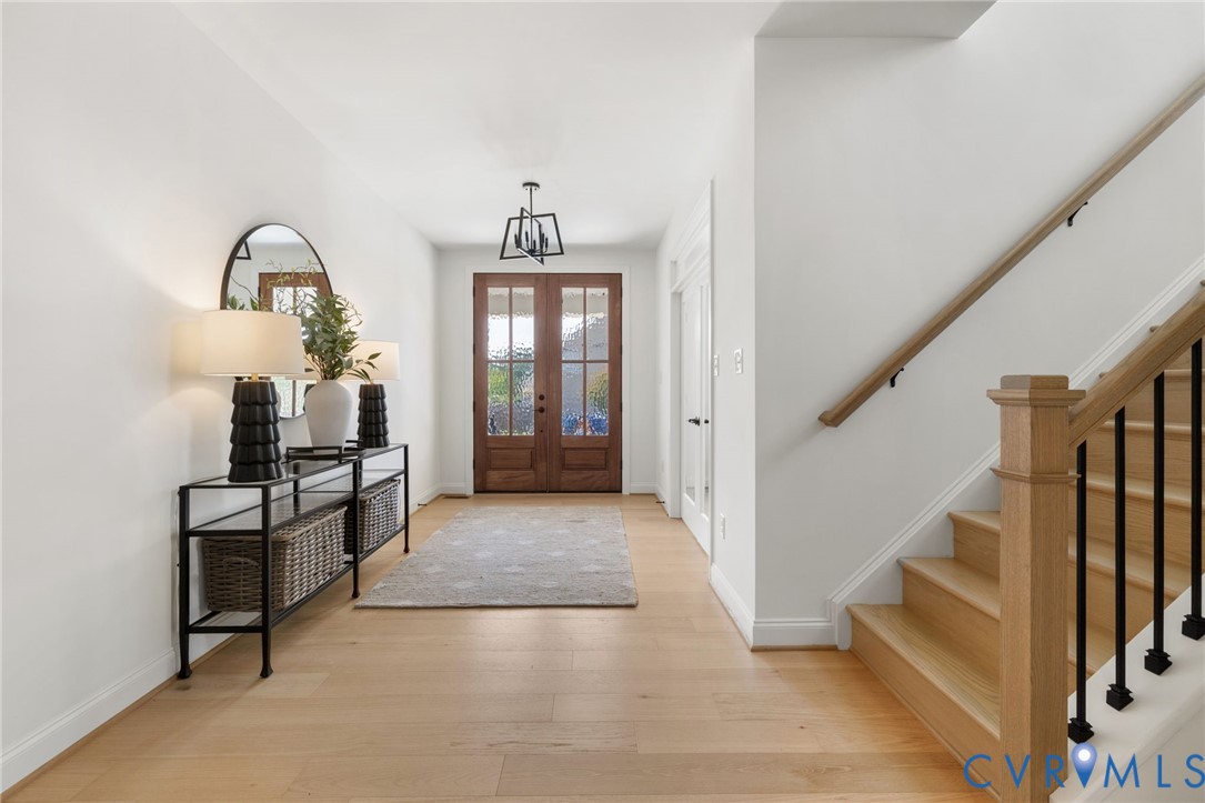 Light wood-finish flooring, white walls, wood double entry doors, wood staircase with black iron spindles, and a modern ceiling fixture