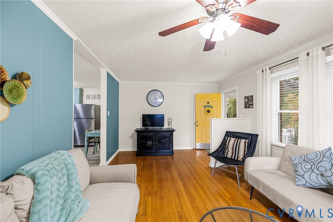 Hardwood flooring, painted accent wall, ceiling fan, multiple windows with blinds, and a brightly colored entry door