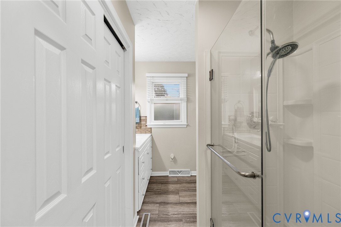 Glass-enclosed shower with a multi-function showerhead, white vanity with multiple drawers, and wood-finish flooring