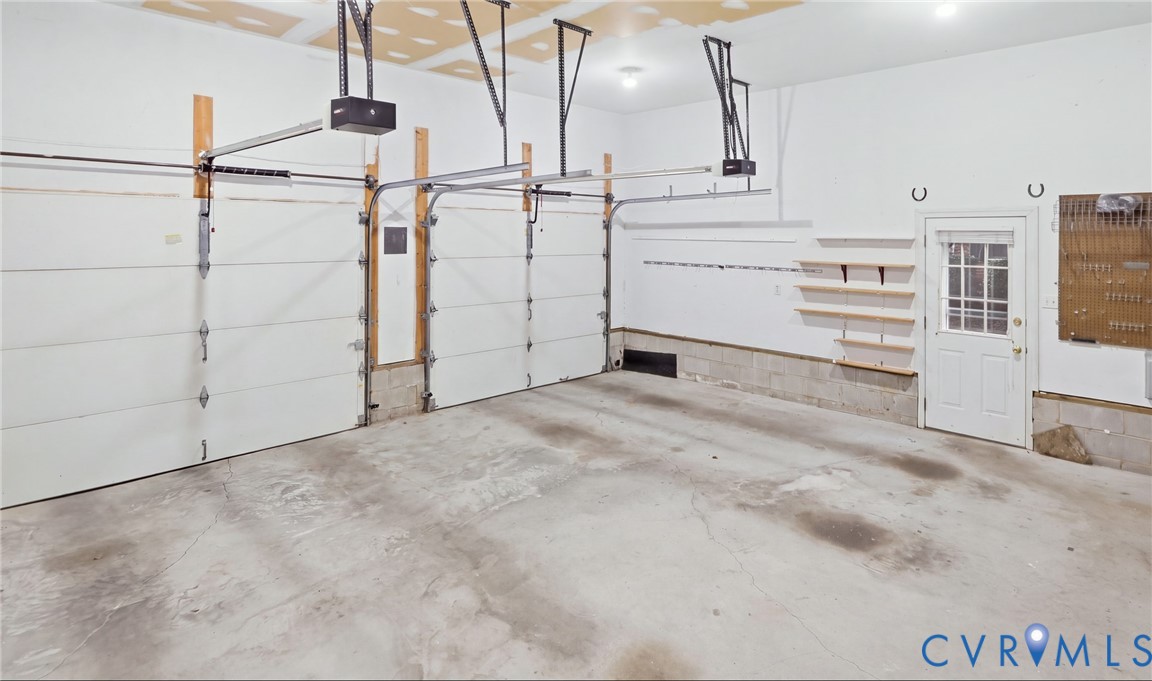 Two overhead garage doors with automatic openers, concrete flooring, overhead lighting, side door access, and wall-mounted shelving