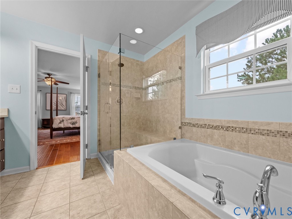 Tiled flooring, built-in soaking tub with deck-mounted faucet, glass-enclosed shower with tiled surround and recessed lighting, and a double-paned window