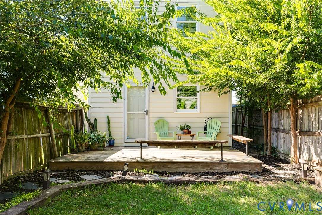 Rear exterior with a wood deck, mature trees, a privacy fence, and a green lawn