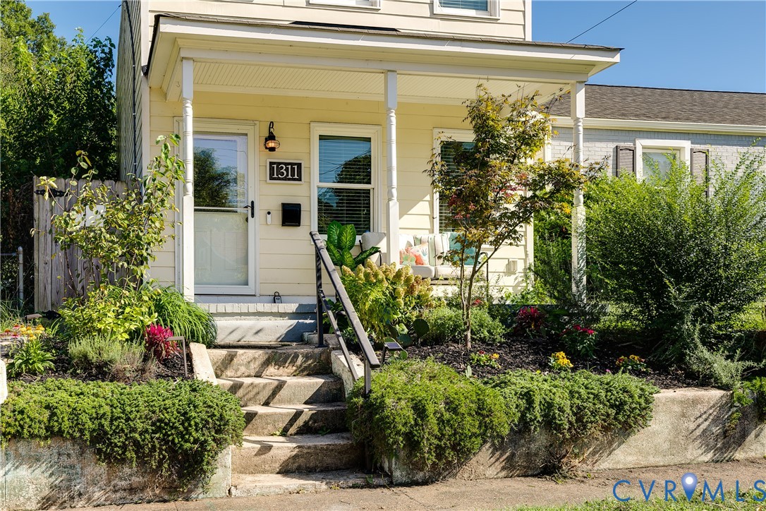 Inviting covered porch with white columns, concrete entry stairs, wrought iron railing, and established landscaping