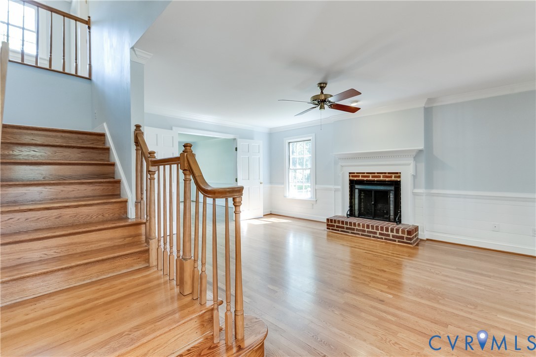 Hardwood flooring throughout, brick fireplace with white mantel, ceiling fan with light fixture, double-hung window, and a wood staircase with balusters