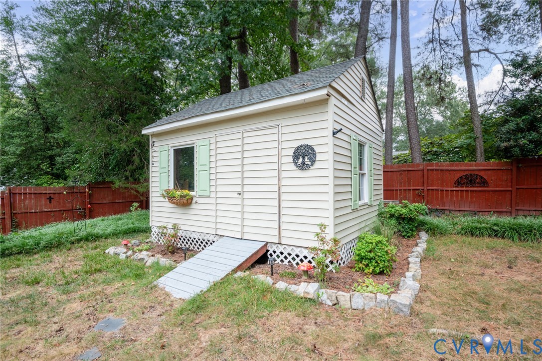 Detached outbuilding with light-colored siding, a green shingle roof, a window with green shutters, and a wood access ramp