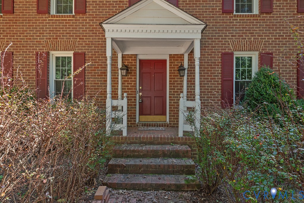 Brick facade with red shutters, featuring a covered porch entry with white columns and railings, and a red paneled front door
