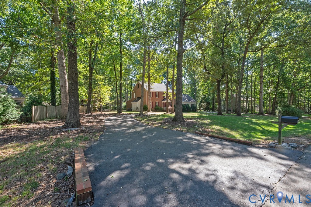 Paved driveway leading to a brick residence with white trim and shuttered windows, surrounded by mature trees and a manicured lawn