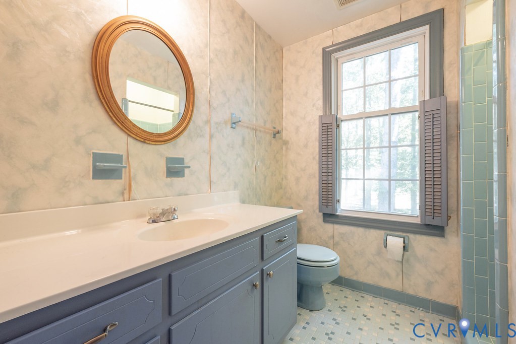 White countertop vanity with built-in sink and blue cabinetry, framed oval mirror, grid window with louvered shutters, and tiled flooring