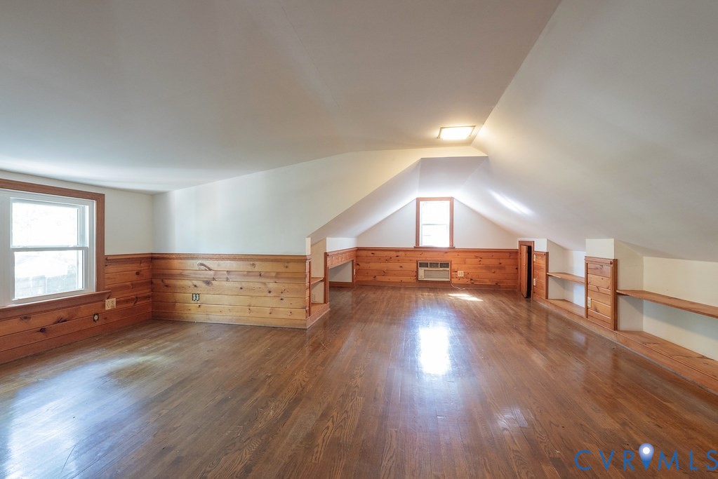Spacious room featuring wood-finish flooring, wood paneling, two windows, built-in shelving, and a sloped ceiling
