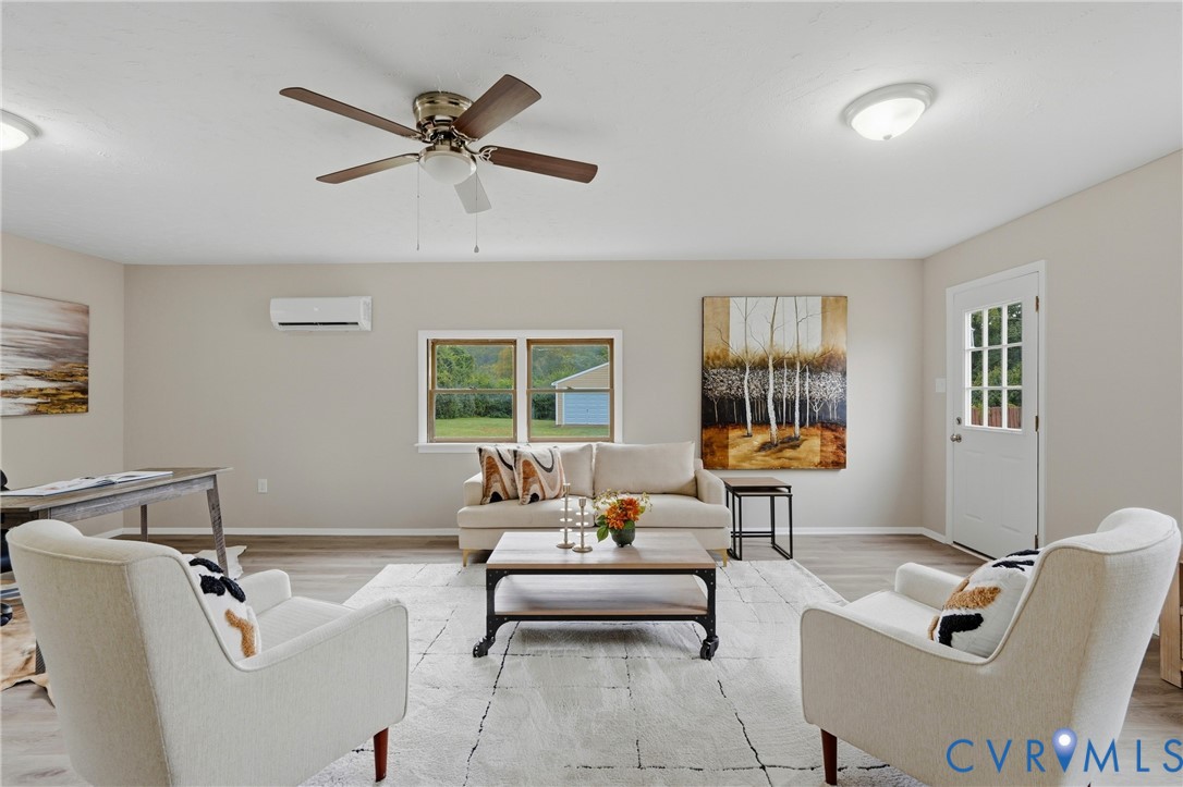 Wood-finish flooring, ceiling fan with integrated lighting, wall-mounted air conditioning unit, multiple windows, and a glass-paneled entry door