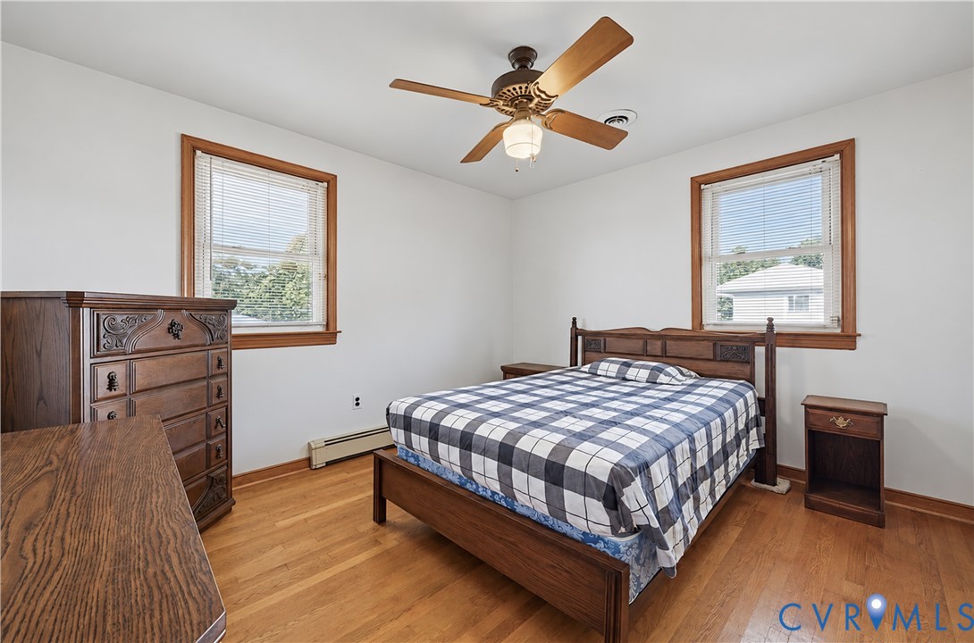 Spacious secondary bedroom featuring hardwood flooring, two large windows with wood-trimmed frames providing natural light, wood-finish built-in dresser, ceiling fan with integrated lighting, and wood-frame bed frame