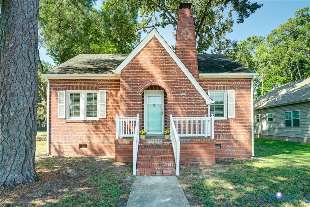 Brick-clad exterior with a prominent chimney, white window shutters, a front porch with white railings, and a light blue entry door