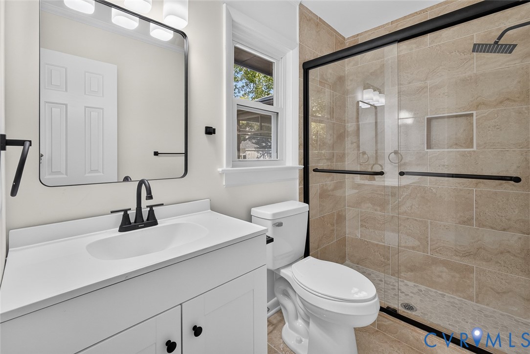 Bathroom featuring a white vanity with an integrated sink, matte black faucet, rectangular mirror, walk-in shower with stone-tile surround, and a glass enclosure with a black frame