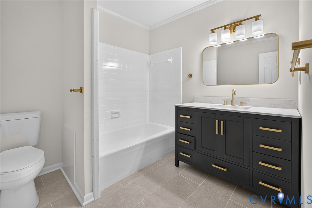 Dark charcoal vanity featuring brass hardware and a white countertop, rectangular vessel sink, wall-mounted brass faucet, arched vanity mirror, and neutral floor tile