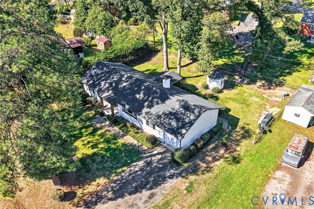 Aerial perspective of a single-story residence featuring an asphalt shingle roof, a central masonry chimney, an attached garage, a paved driveway, and mature landscaping