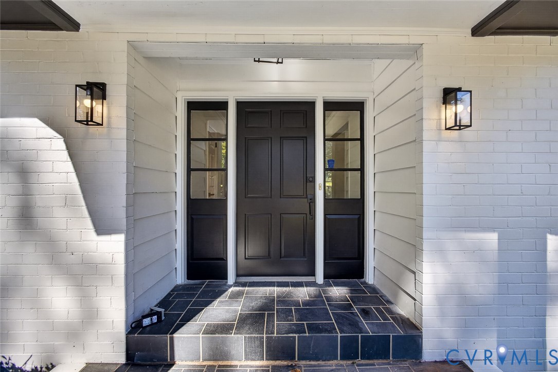 Entryway featuring painted brick exterior, six-panel dark finish door, flanking glass-paneled sidelights, geometric tile flooring, and rectangular wall-mounted lanterns
