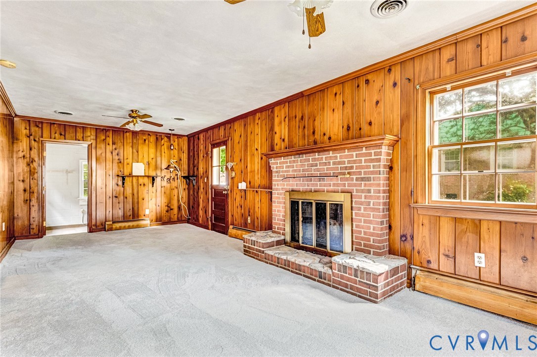 Brick fireplace with hearth, wood-paneled walls, carpet flooring, window with exterior exposure, and ceiling fan
