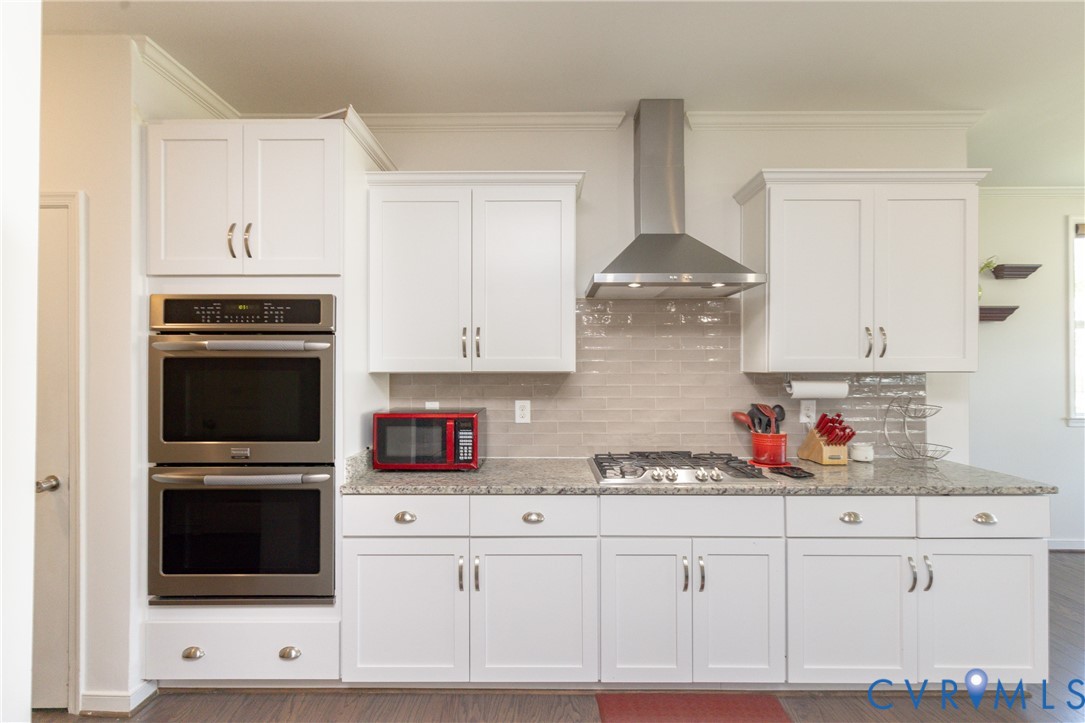White shaker-style cabinetry, stainless steel double wall ovens, gas range with stainless steel hood, light-toned tile backsplash, and light-toned speckled countertops