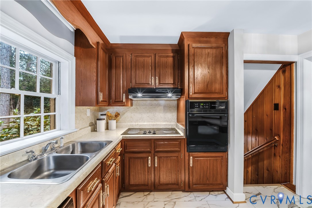 Wood cabinetry with brass hardware, double basin stainless steel sink, built-in wall oven, electric cooktop, and white tiled flooring Wood cabinetry with brass hardware, double basin stainless steel sink, built-in wall oven, electric cooktop, and white tiled flooring