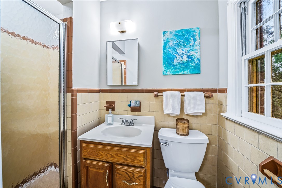 Frosted glass shower enclosure, wood-finish vanity with white countertop, recessed medicine cabinet, wall-mounted light fixture, and tile wainscoting