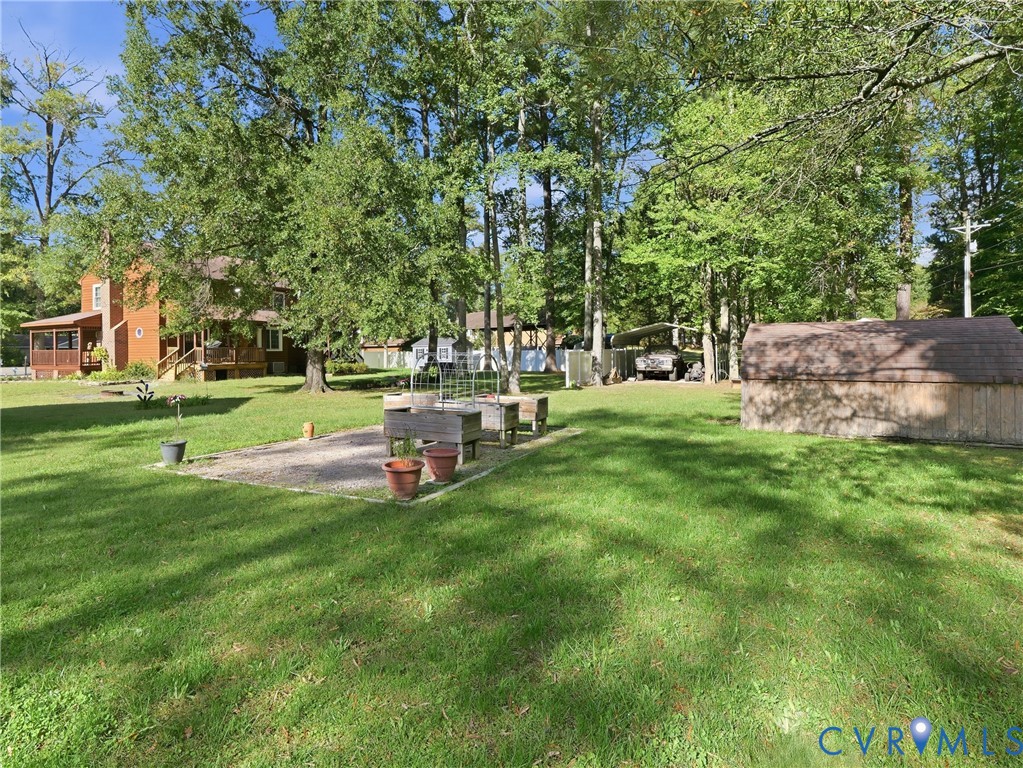 Expansive lawn featuring mature trees, a gravel-based garden area with raised wood planter boxes, a detached storage structure, and a covered parking area
